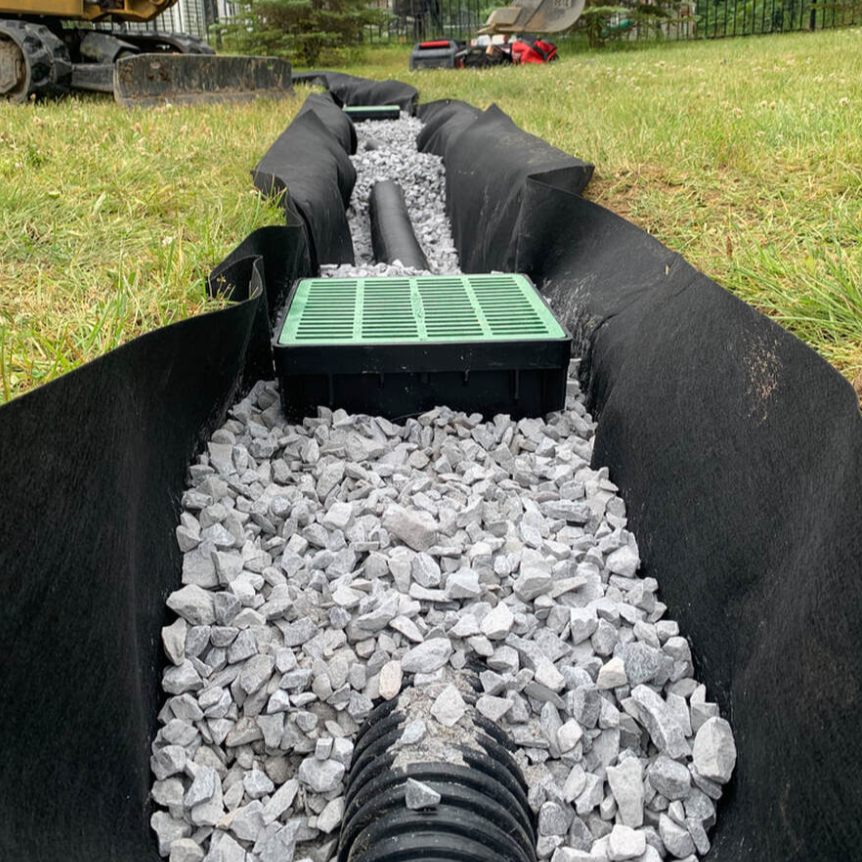 A construction or landscaping trench lined with black fabric, filled with rocks, with pipes and a green crate inside, in a grassy outdoor area.