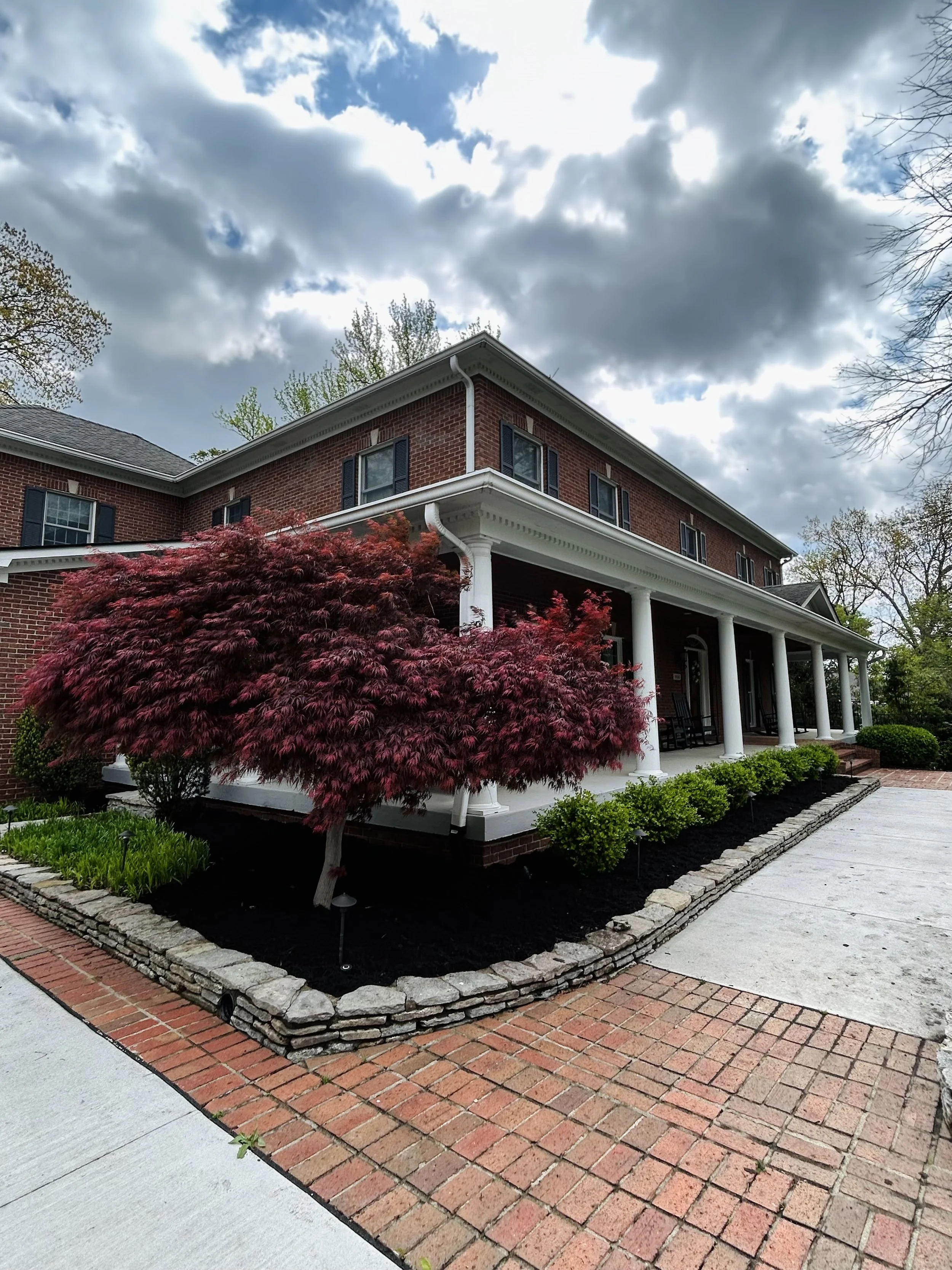 A two-story red brick house with white columns and black shutters, surrounded by landscaped bushes and a red-leafed tree in front, under a partly cloudy sky.
