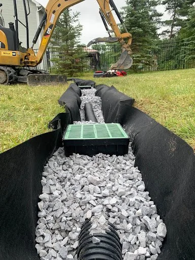 Underground drainage or utility pipe installation with gravel, black fabric liner, and construction equipment in a backyard