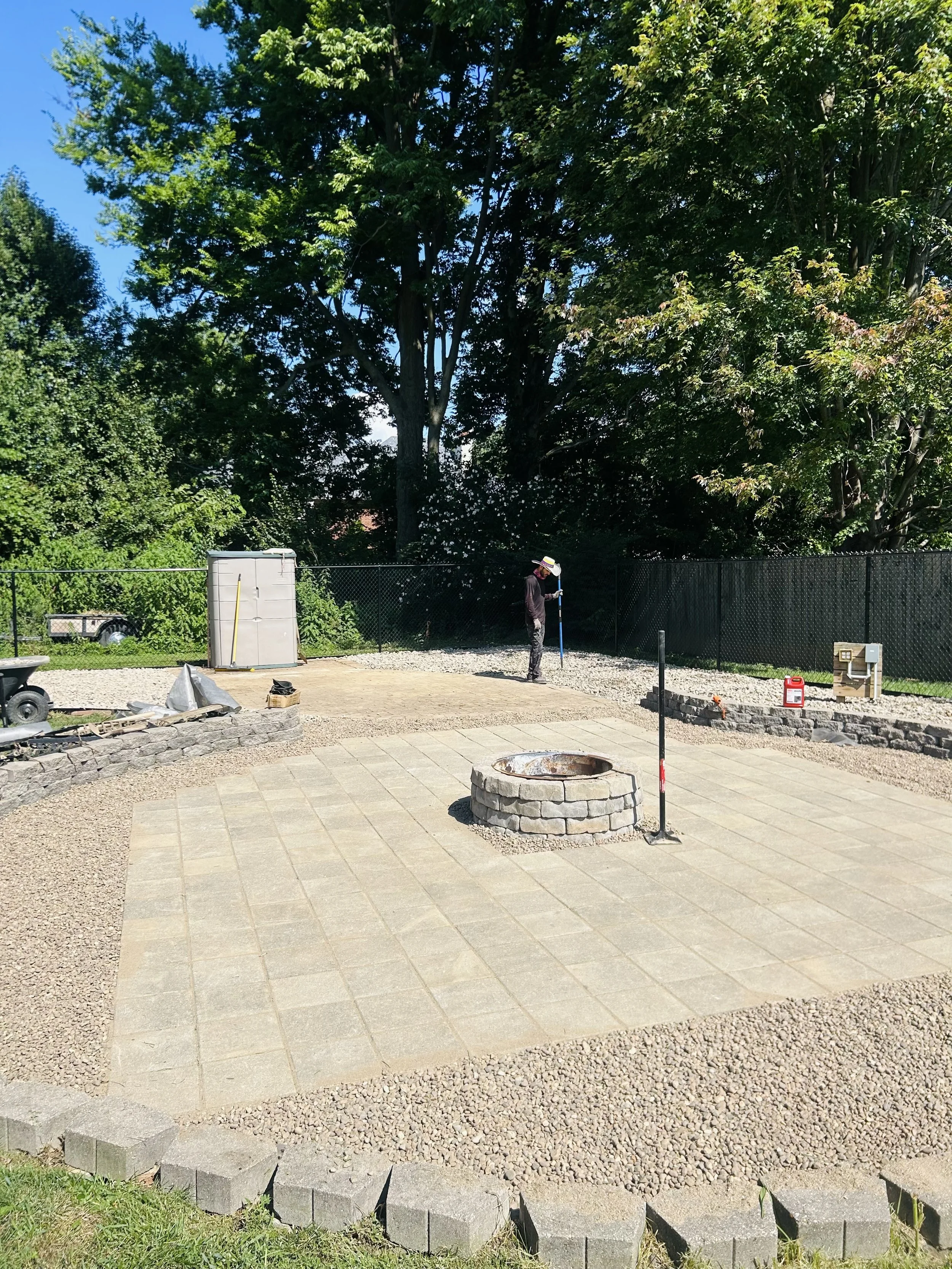 A person is working on a backyard patio. The area has newly laid paving stones and a central fire pit made of bricks. There are construction tools and materials around, including a weed barrier fabric, a level, and small piles of gravel. The backyard is enclosed by a black chain-link fence, with large trees and green foliage in the background under a clear blue sky.