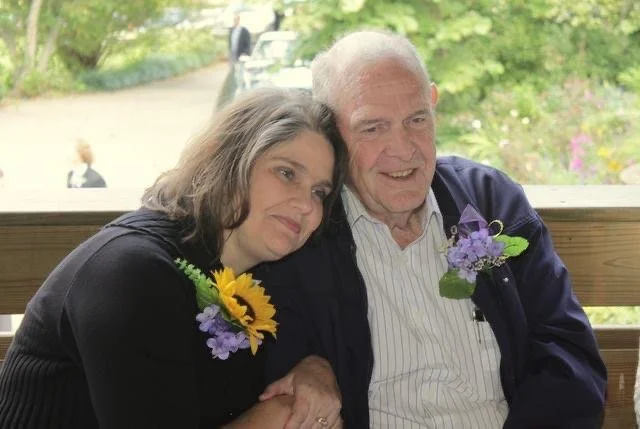 A woman and her father sit close together on a wooden porch,  smiling and enjoying a moment together.