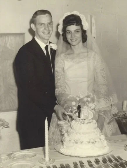 A black and white photo of a newlywed couple cutting a wedding cake. The groom is wearing a dark suit and tie, and the bride is wearing a lace wedding dress with a veil and floral headpiece.