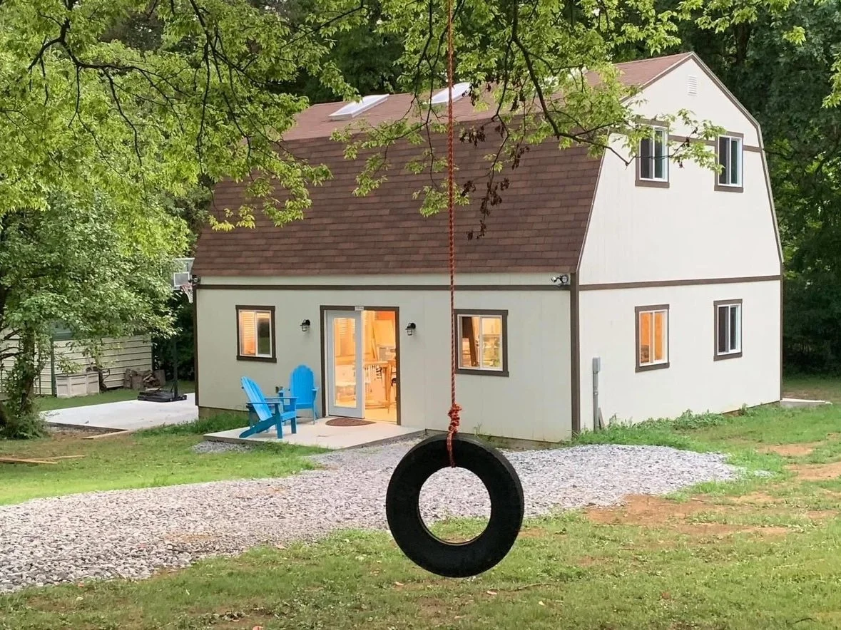 A two-story white barn, surrounded by green trees. A black tire swing hangs from a tree branch in the foreground. The house has several windows and French doors