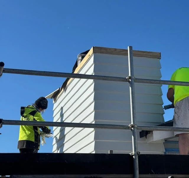 Workers in yellow safety shirts installing or repairing a white exterior wall or siding on a building, with a clear blue sky in the background.