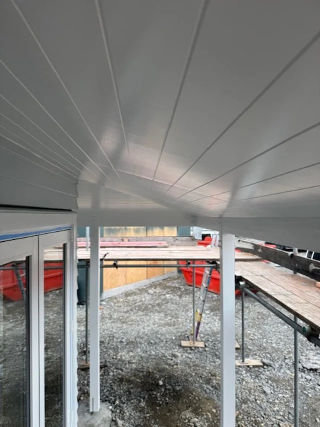 View of the underside of a building with white siding, showing support beams and a partially visible sliding glass door on the left, with construction materials and gravel ground outside.