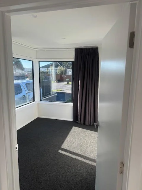 Empty room with white walls, dark carpet, large window with black curtains, outside view through the window showing a car and trees.