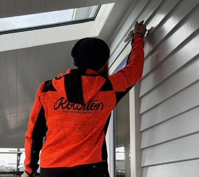 A person wearing an orange and black jacket with 'Rowton Painting & Decorating Ltd.' on the back, painting or touching up the exterior wall of a house.