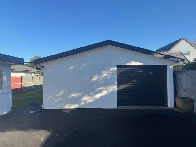 White garage with black roll-up door in a residential backyard under a clear blue sky.