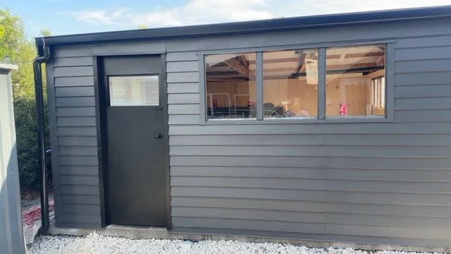 Exterior of a gray shed with a small window and a door, situated on a gravel surface with trees in the background.