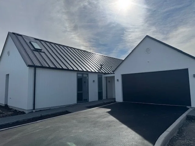 A modern white house with a metal roof, two front doors, and a garage with a black door, under a partly cloudy sky.