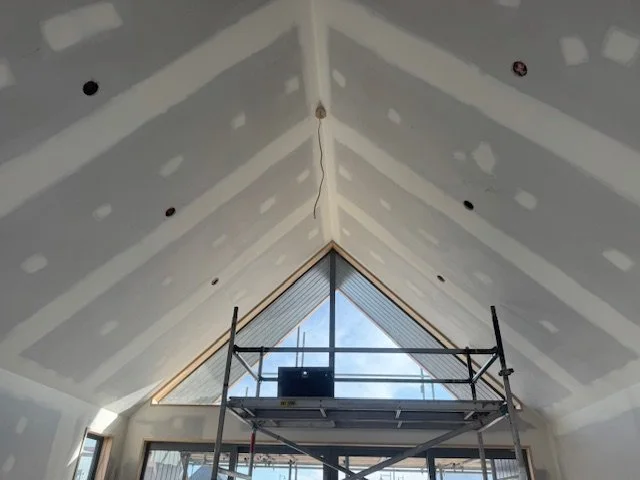 Interior view of a room under construction with finished drywall ceiling and a large triangular window at the front, exposing blue sky and clouds, with scaffolding inside.