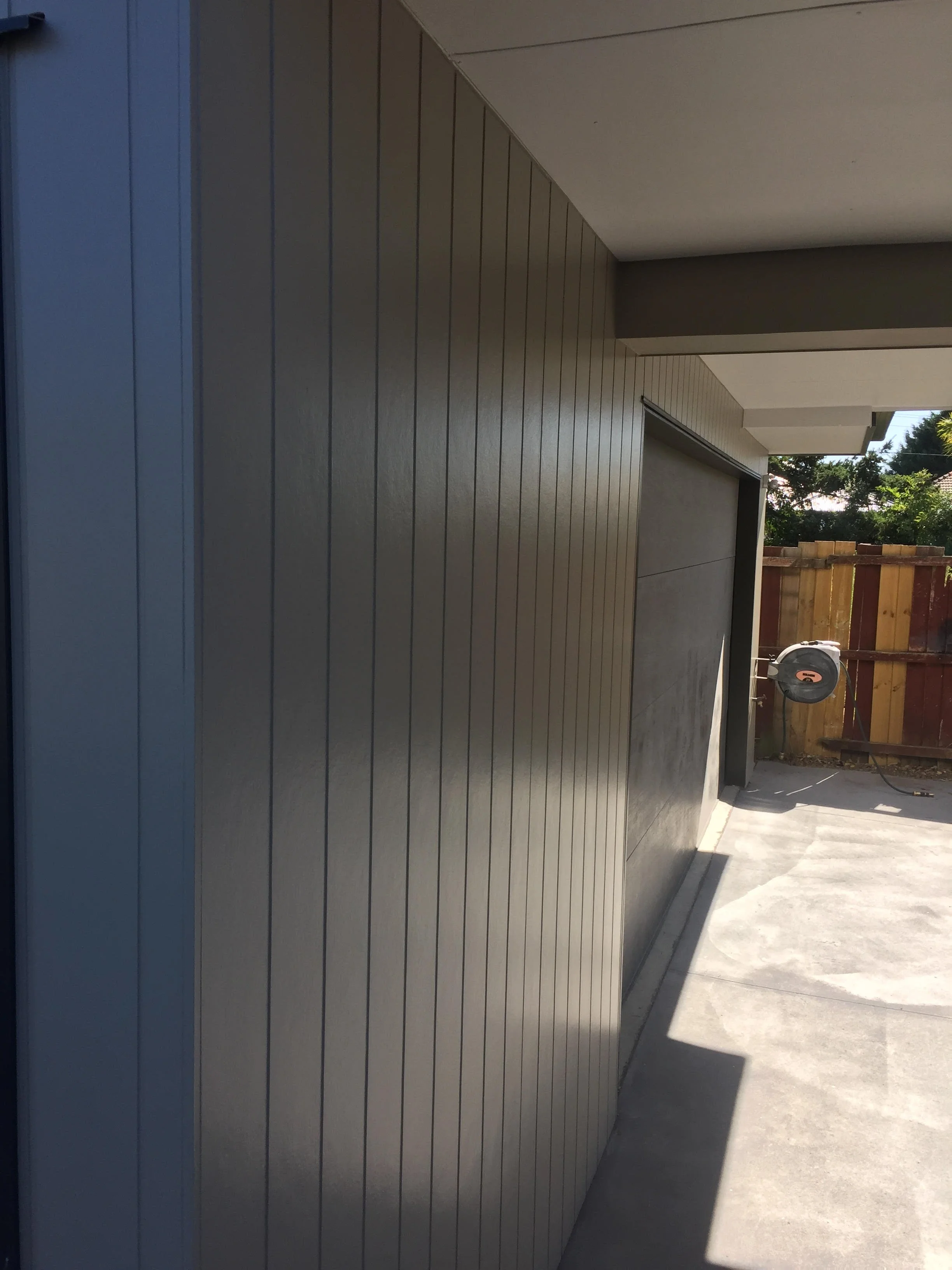 Side view of a modern garage with a closed gray door, adjacent to a concrete driveway, and a hose reel mounted on a wooden fence in the background.
