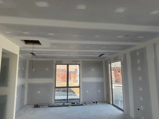 Interior of a room under construction with drywall panels installed, exposed ceiling fixtures, and sliding glass doors leading outside where a wooden fence is visible.