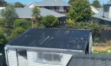 The image shows the roof of a house with a black corrugated metal roof and neighboring houses with trees in the background.