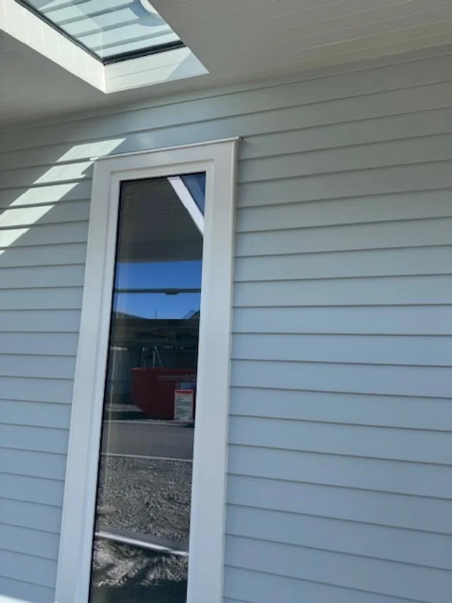 Close-up of a house's exterior corner with white siding, a tall window, and a porch roof with a skylight.