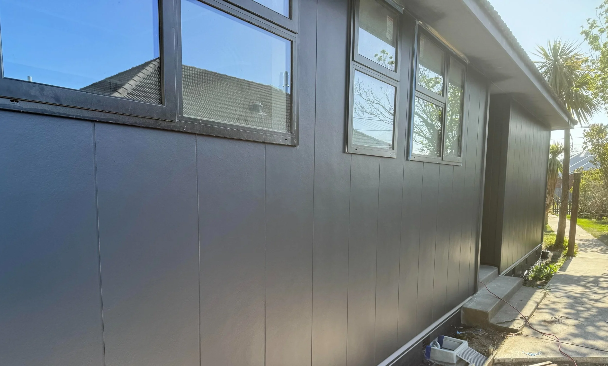 Side of a house with black siding and multiple windows, some of which are reflecting the sky and nearby roof. The house is on a sidewalk with plants and trees nearby.