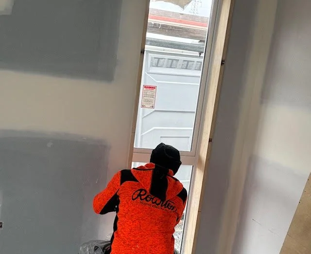 Person wearing an orange and black jacket and black hood, kneeling by a small window in a construction or renovation site.