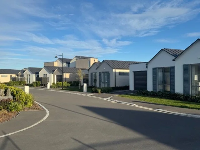 Residential neighborhood with modern white houses, neatly landscaped yards, and a curved street under a blue sky.