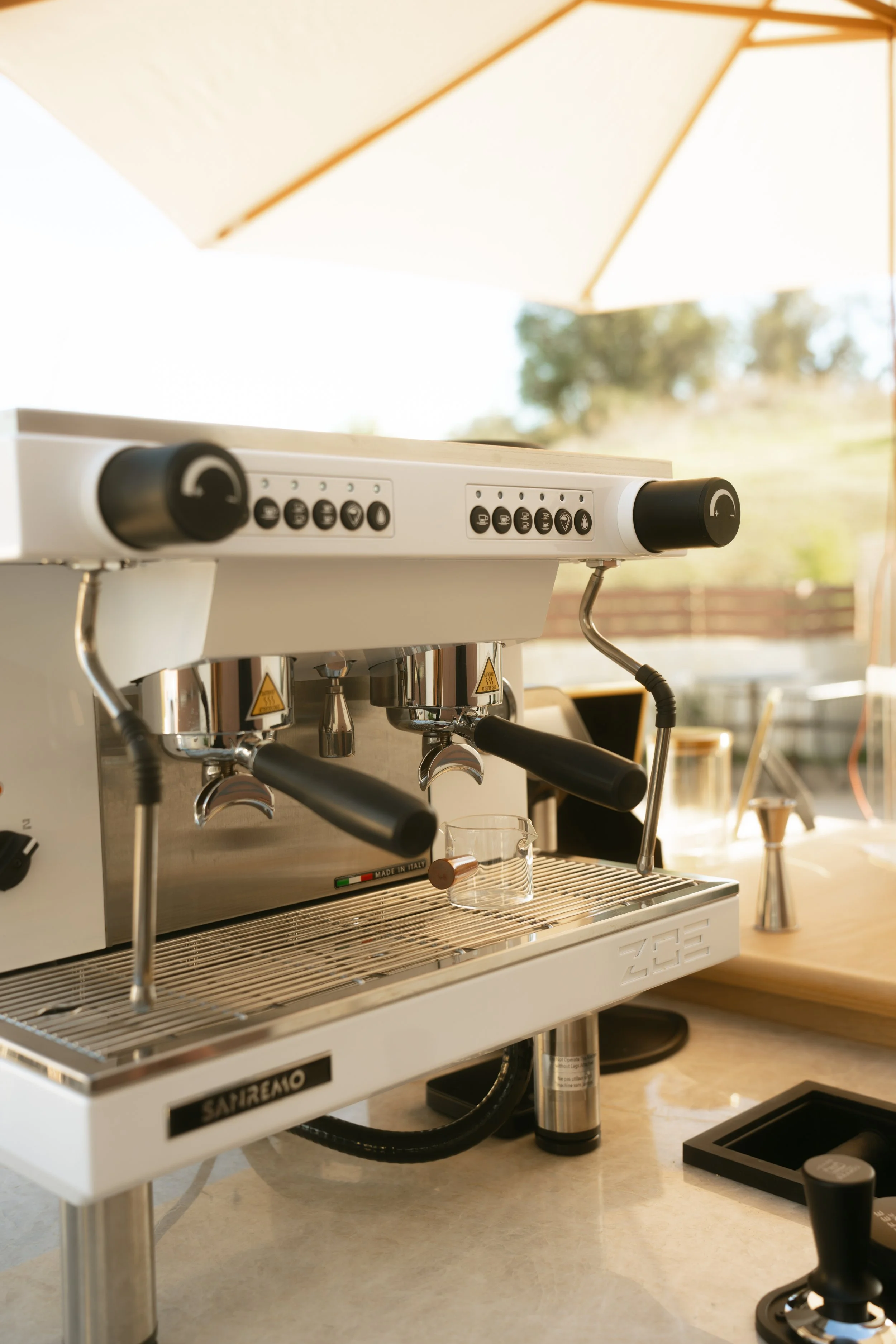 A commercial espresso machine with dual group heads, placed on a countertop outdoors, under an off-white umbrella.
