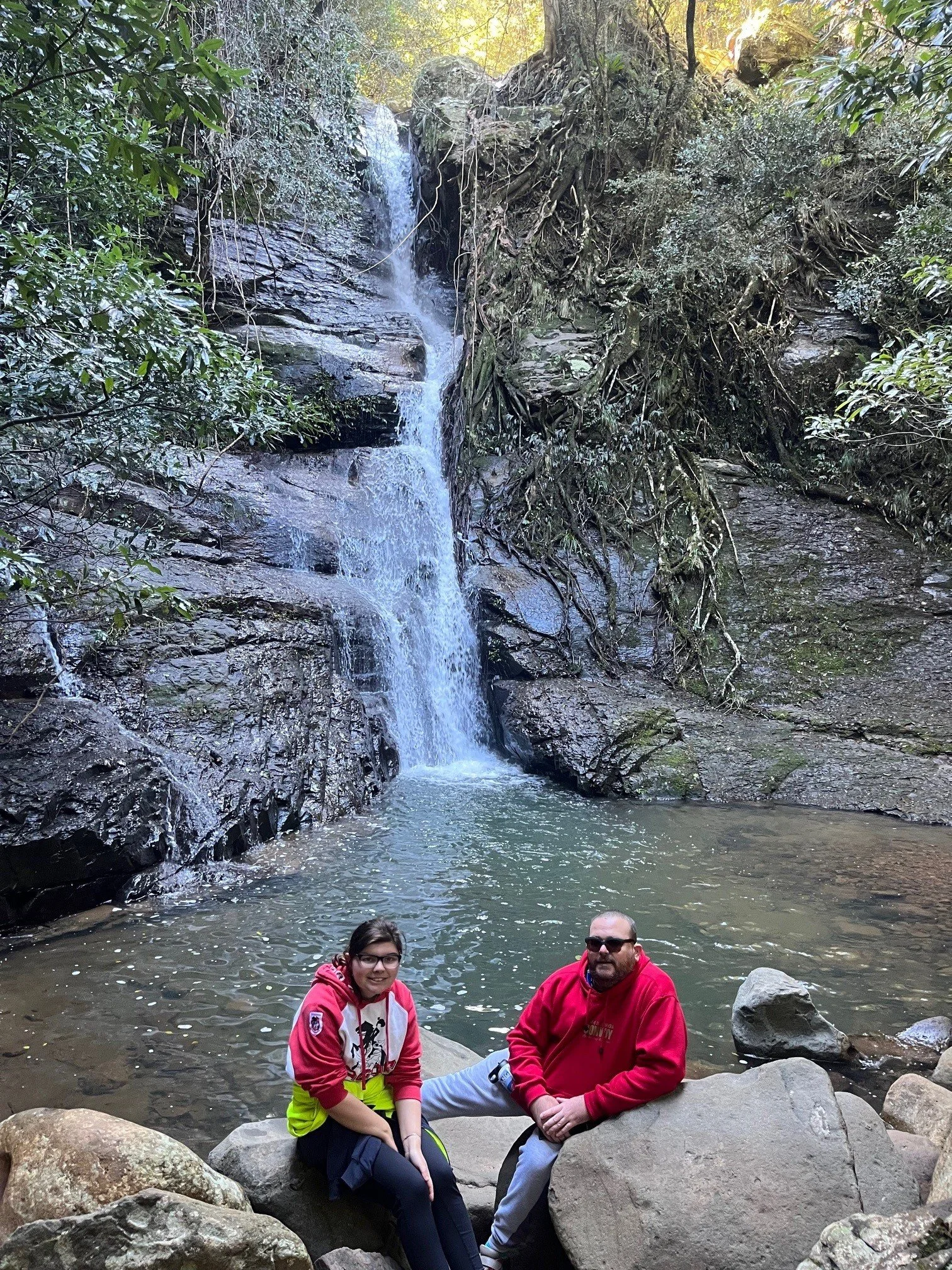 A photo of a man and woman resting on big boulders at the base of a small waterfall