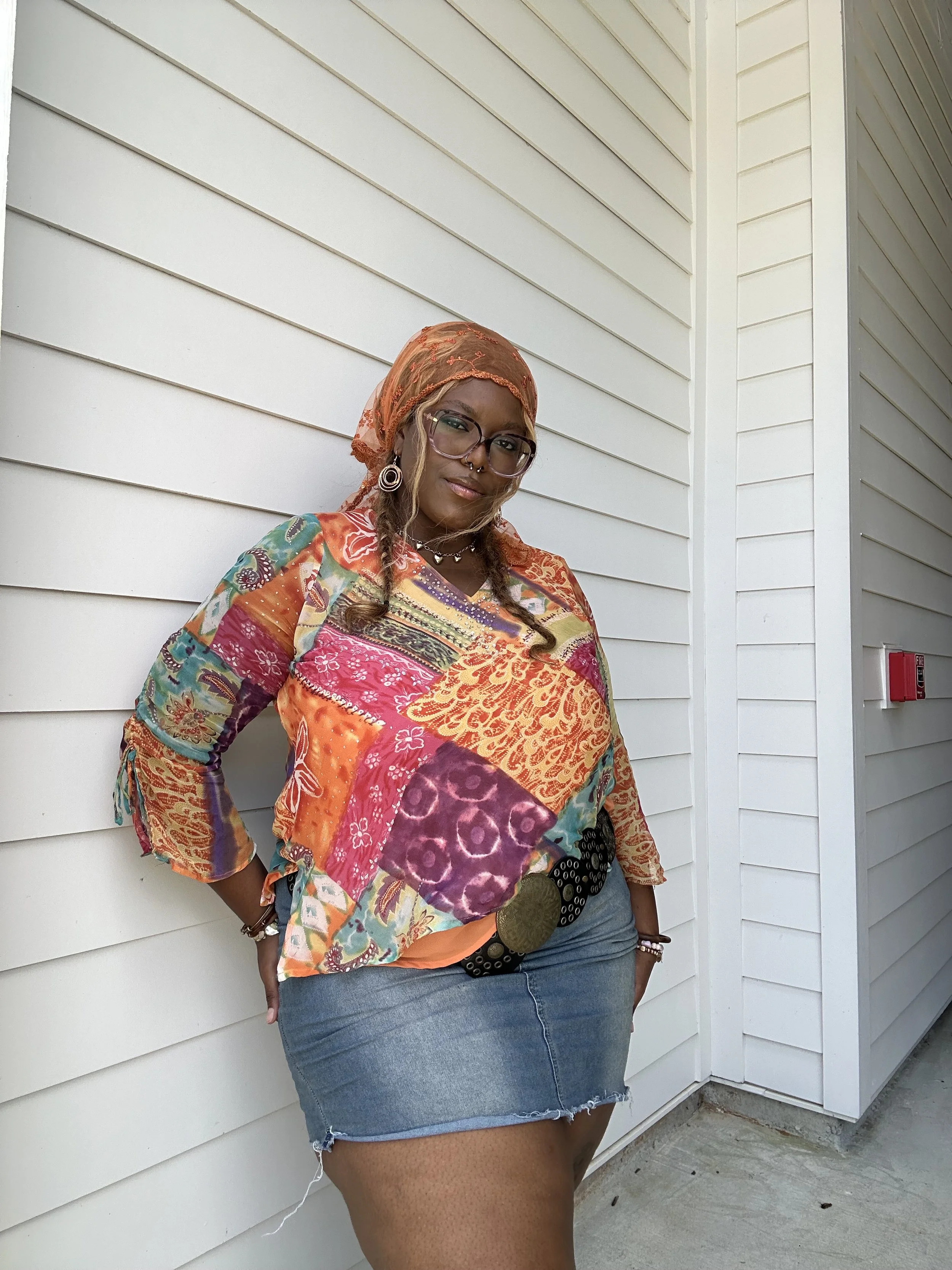 Woman wearing glasses, earrings, a colorful patchwork shirt, denim skirt, standing against a white siding wall with her hand on her hip.
