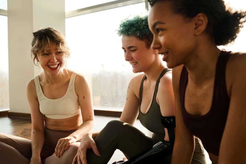 Three women sitting together indoors, smiling and laughing during a casual conversation.