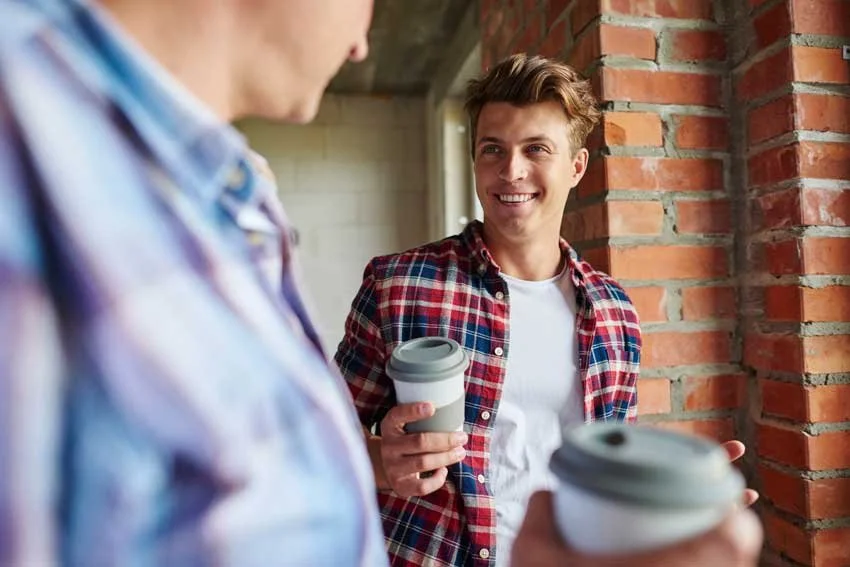 Two young men standing outdoors near a brick wall, smiling and holding coffee cups, engaged in conversation.