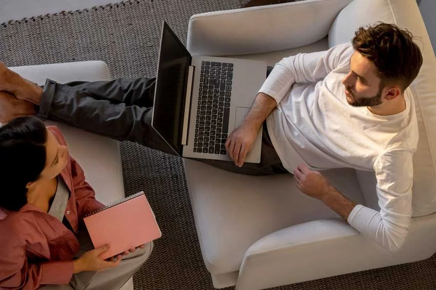 A man resting on a sofa with a laptop on his lap, talking to a woman sitting nearby holding a pink notebook.