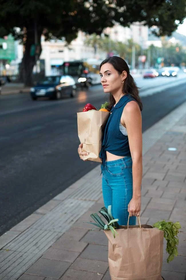 A woman standing on a city sidewalk holding two brown paper grocery bags, one filled with fresh vegetables and produce, and the other with leafy greens and pineapple, with a busy street and cars in the background.