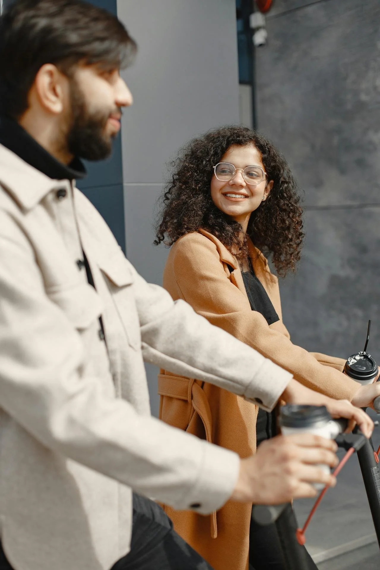 Two people riding electric scooters indoors, smiling and holding coffee cups.