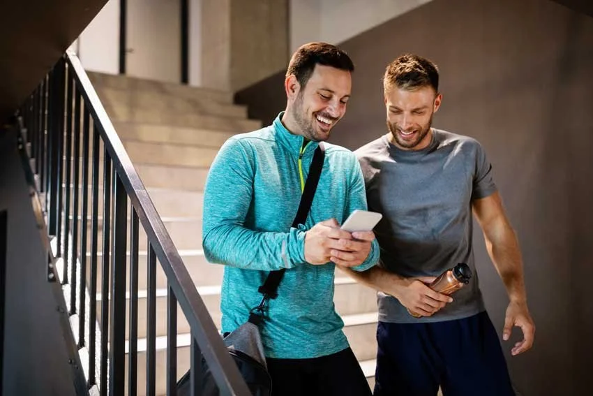 Two young men standing on a staircase, smiling and looking at a smartphone. One is holding a water bottle, the other has a gym bag.