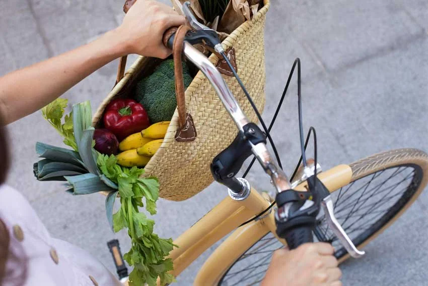 Person riding a yellow bicycle with a woven basket filled with fresh produce like red peppers, bananas, lettuce, and other vegetables.