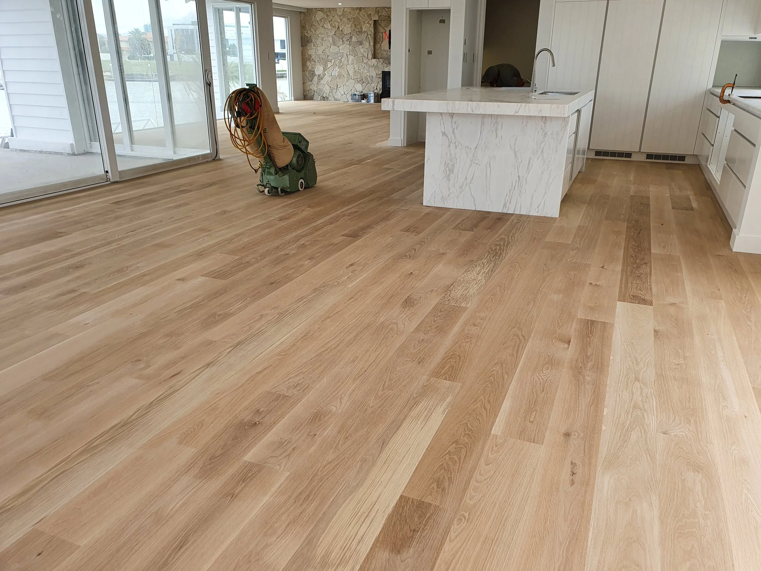 Empty kitchen with newly installed light hardwood floors, white marble kitchen island, and sliding glass doors leading to a patio.