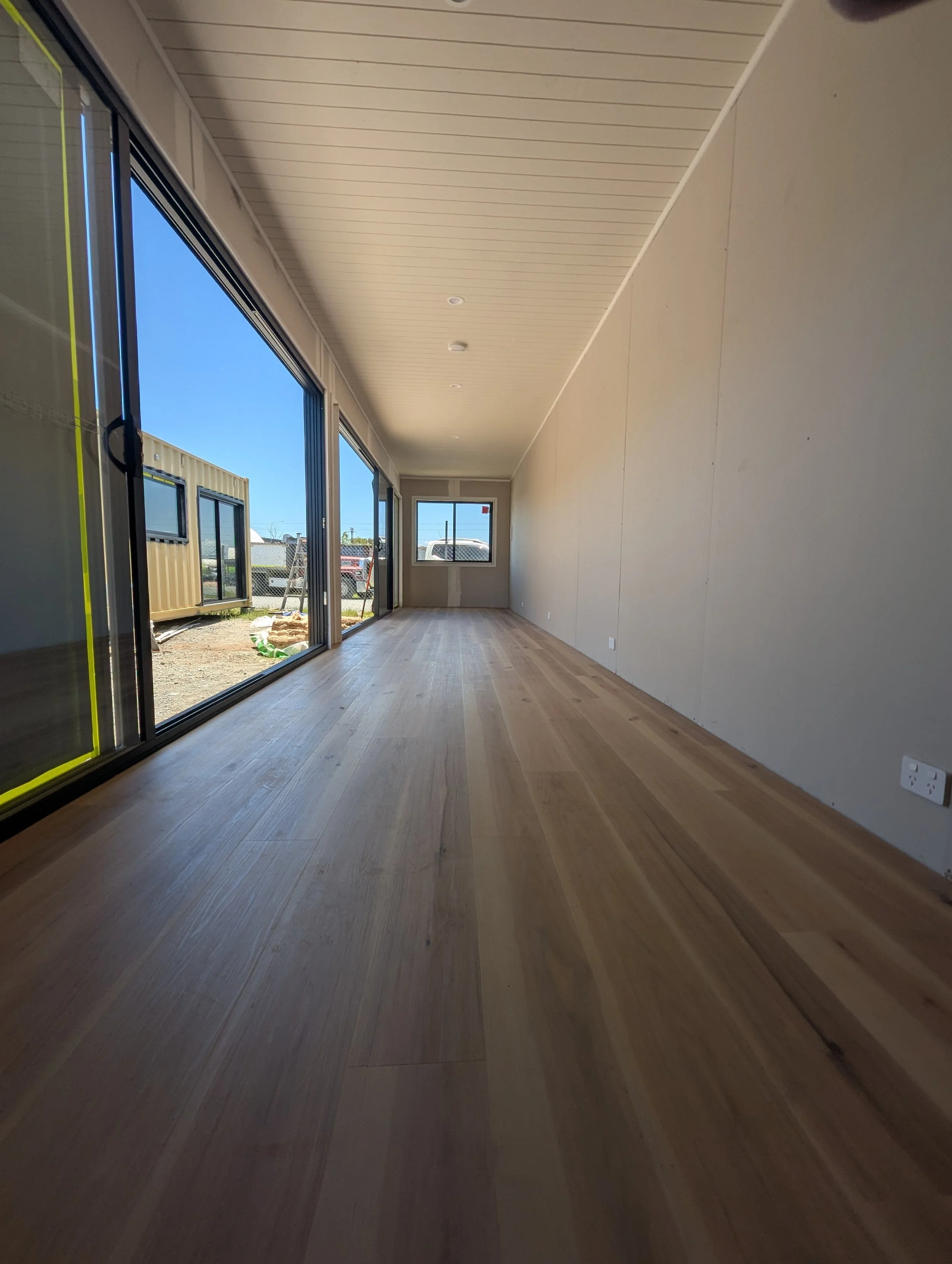 Empty indoor space with wooden floor, large glass sliding doors on the left, and a small window at the far end, under a beige ceiling with recessed lighting.