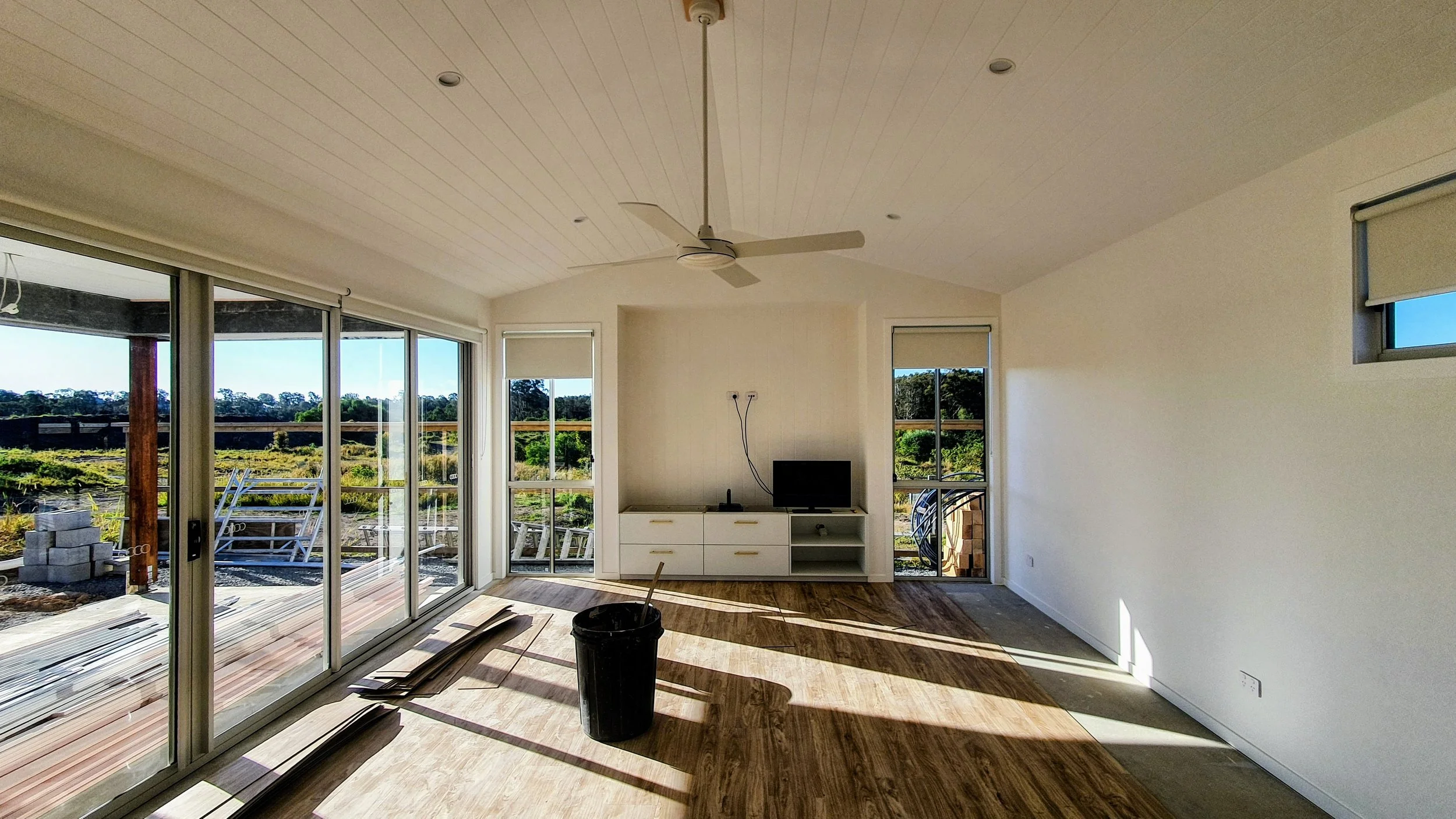 Empty living room with large sliding glass doors, white walls, wooden flooring, a ceiling fan, and windows showing an outdoor view of greenery.