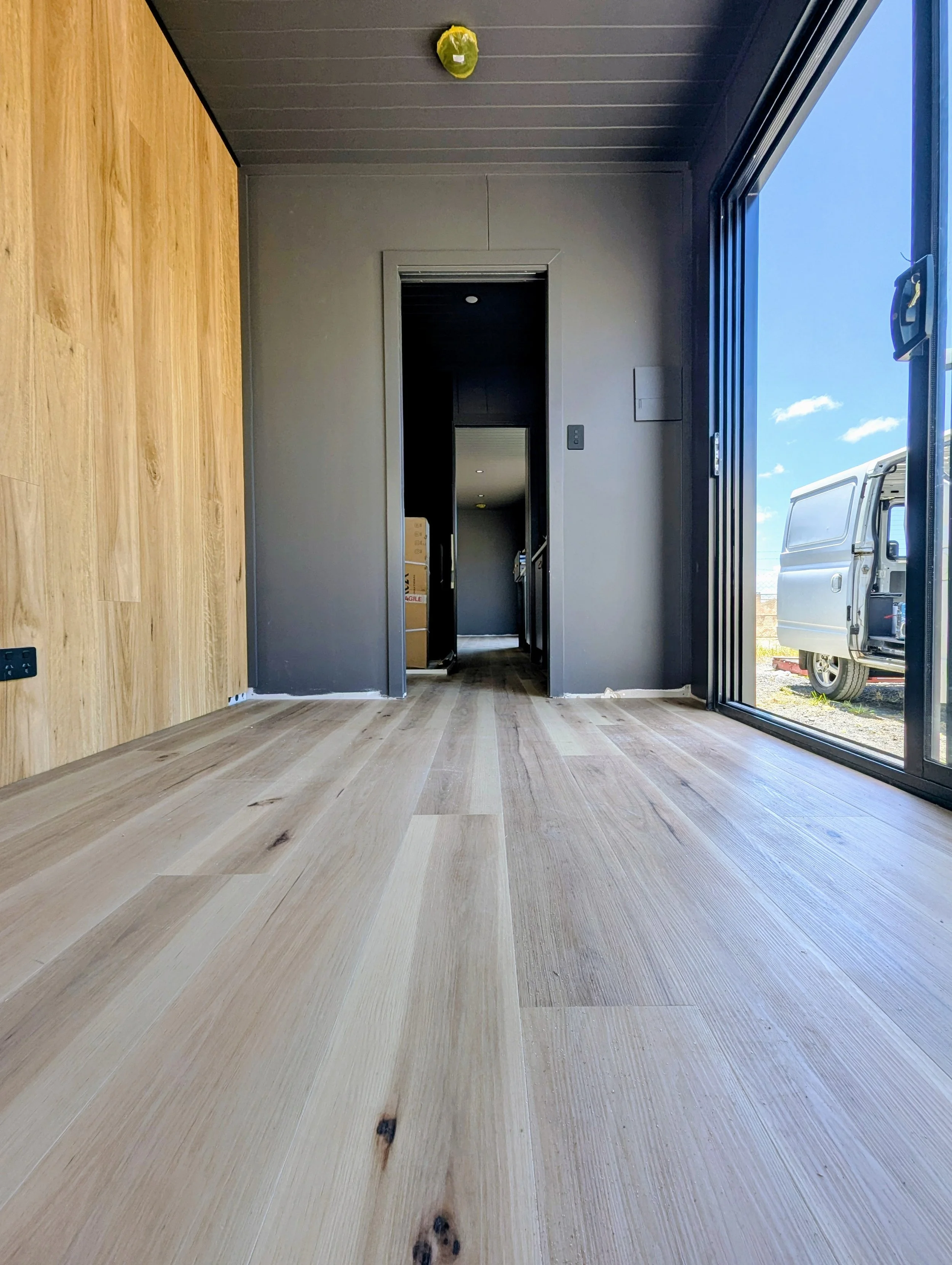 View of a room with wooden floors, gray walls, and a large sliding glass door to the right, with a van parked outside. A small yellow object is on the ceiling.