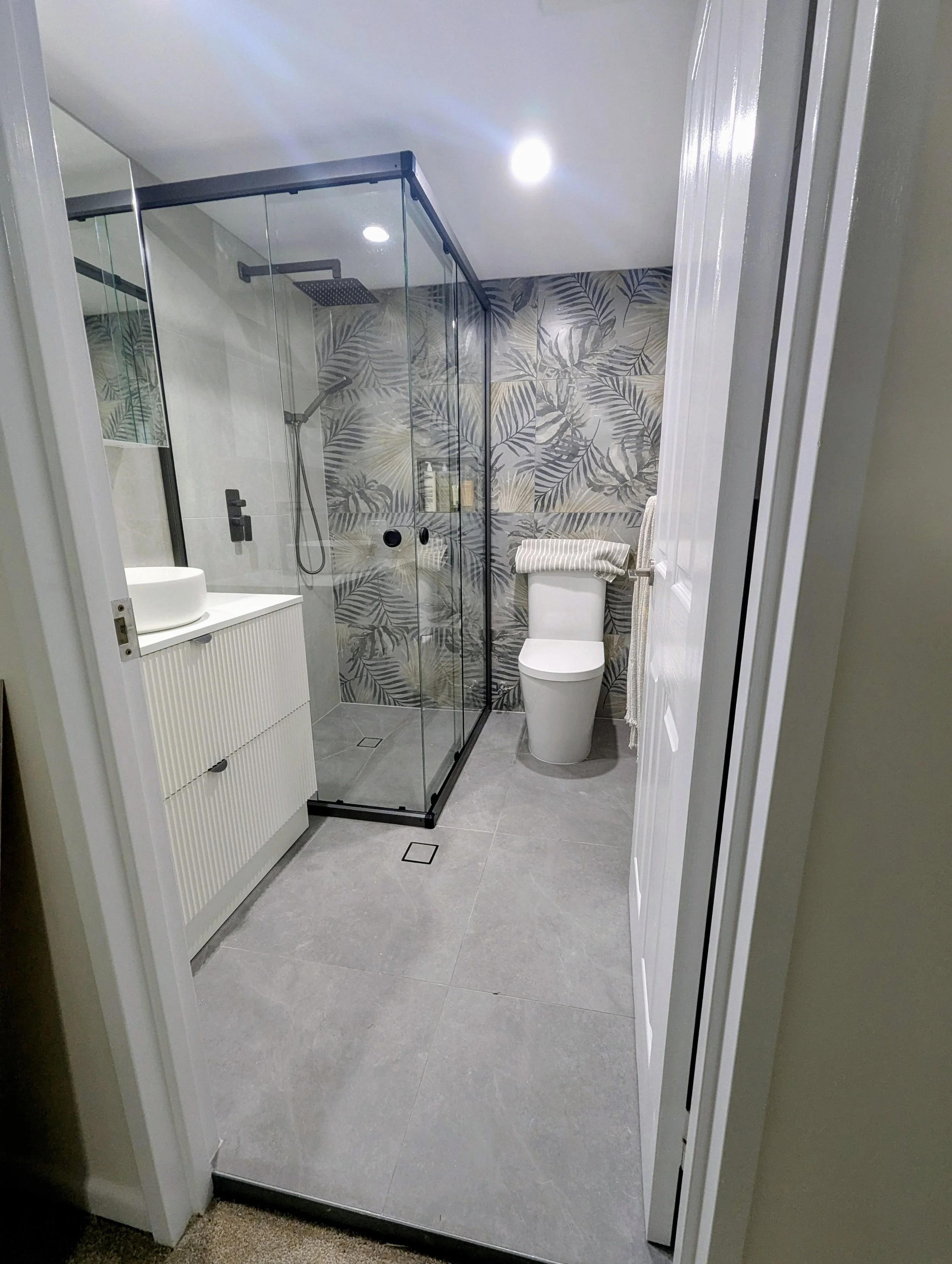 Modern bathroom with a glass-enclosed shower, a white toilet with a striped towel, a white vanity with a round vessel sink, and a wall with botanical wallpaper.