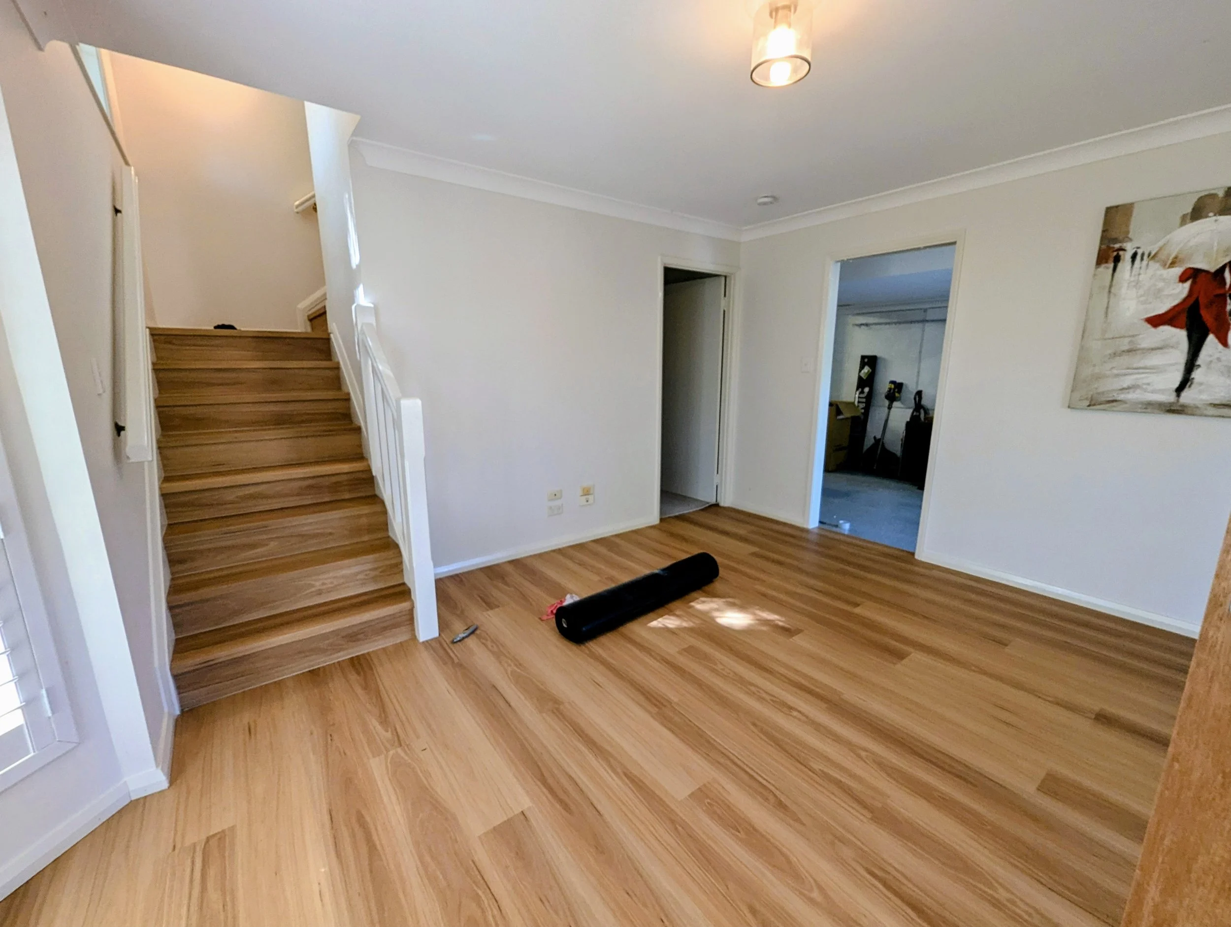 Empty living room with wooden flooring, a staircase on the left, an open doorway on the right, and a painted wall with artwork. A black roller and a utility knife are on the floor.