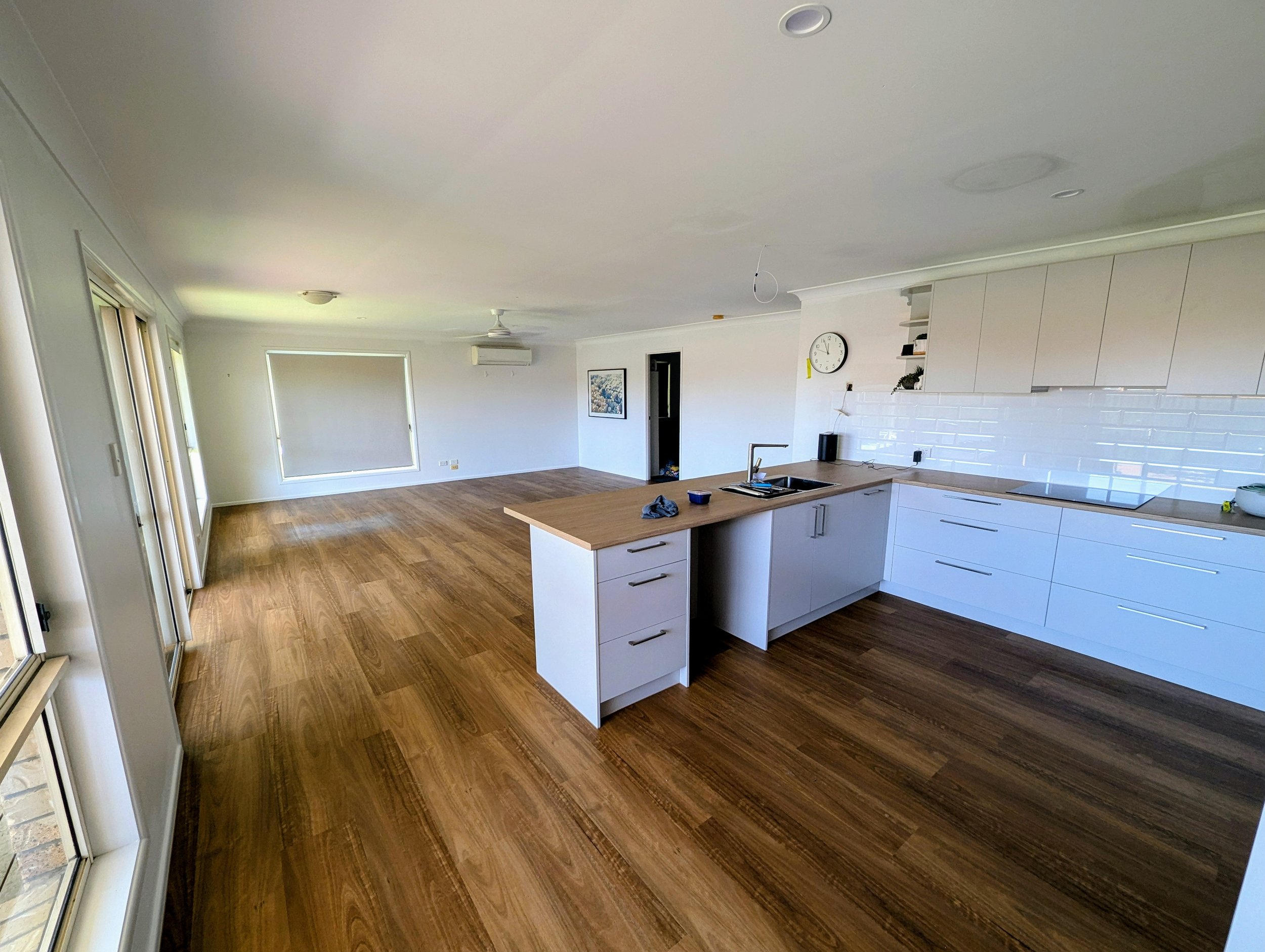 Empty modern open-plan kitchen and living room with wooden floors, white cabinets, and large windows.