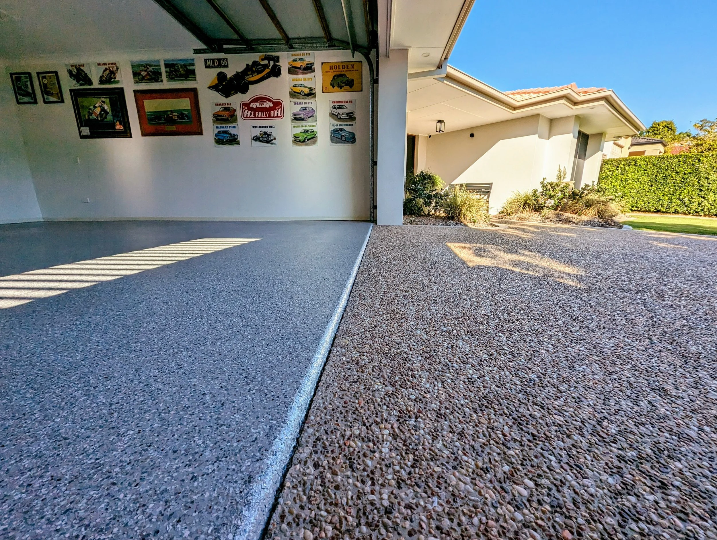 View of a garage with a smooth concrete floor on the left and textured stained concrete on the right, opening to a landscaped yard with plants and a hedge, under a clear blue sky.