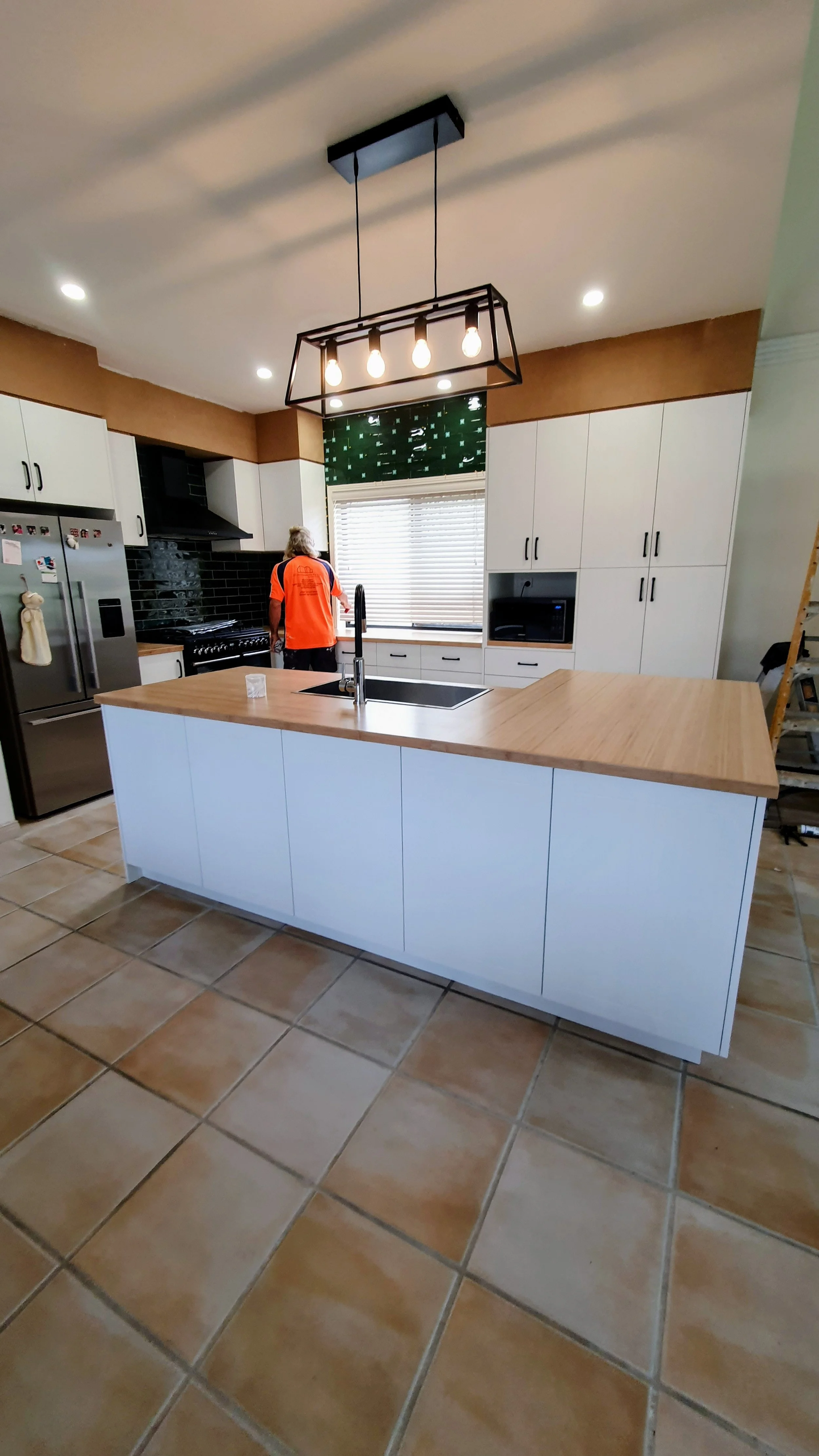 Modern kitchen with a white island and wooden countertop, white cabinets, black backsplash, stainless steel refrigerator, and a man in an orange shirt near the window.