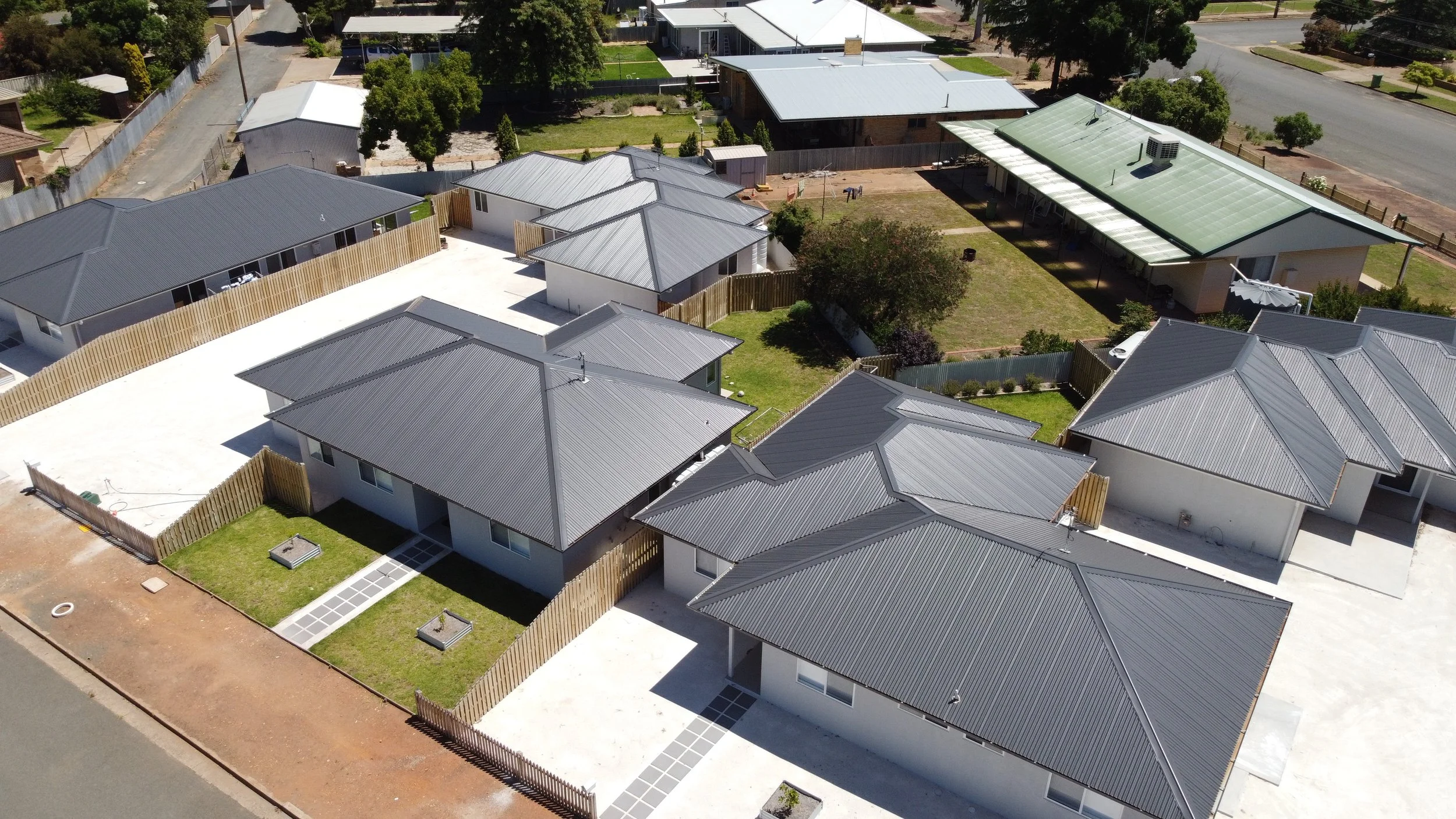 Aerial view of a residential neighborhood with multiple modern houses with gray metal roofs, fenced yards, and paved walkways in a suburban setting.