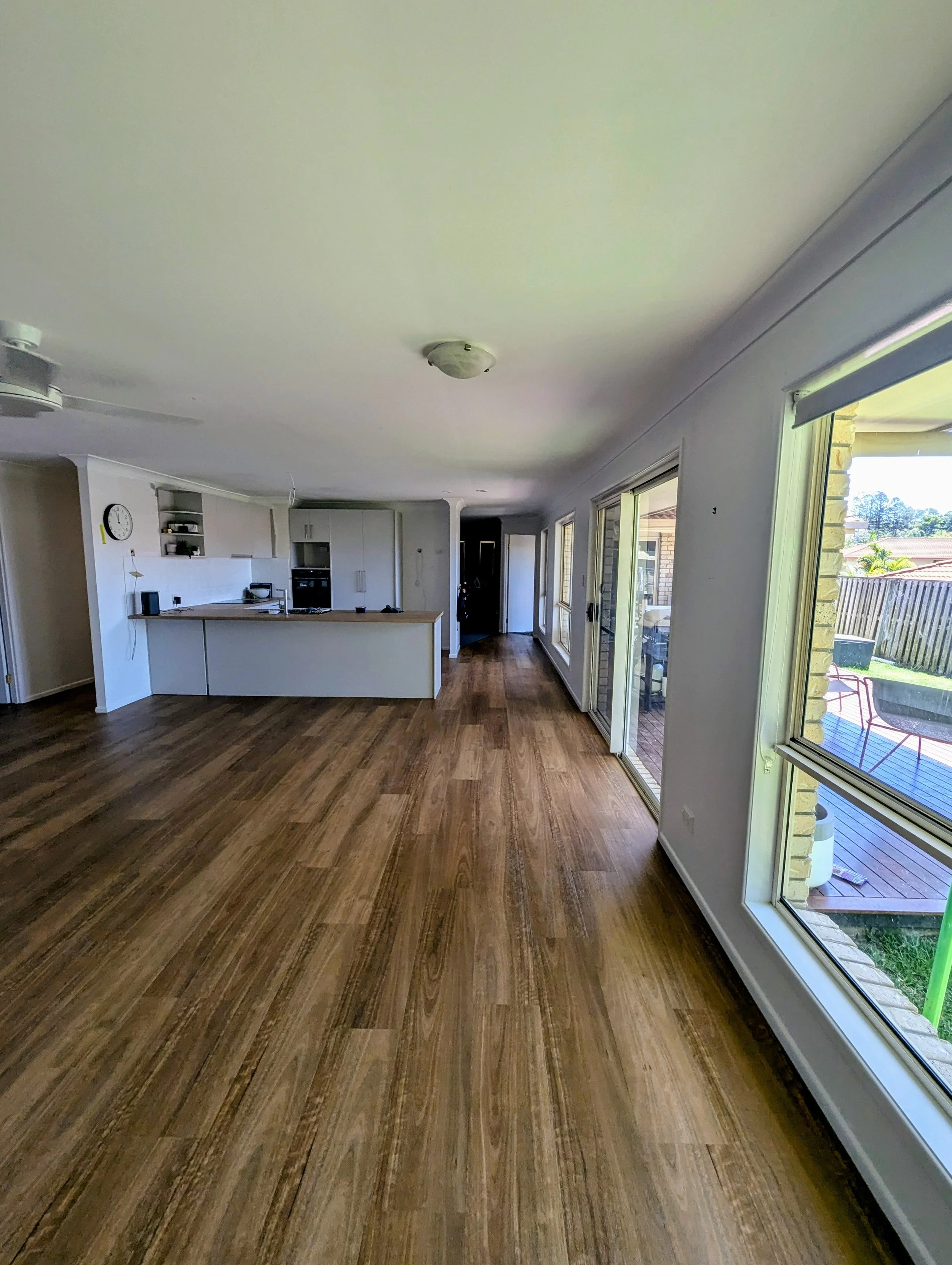 Empty living area with large windows, wooden floor, and kitchen in the background