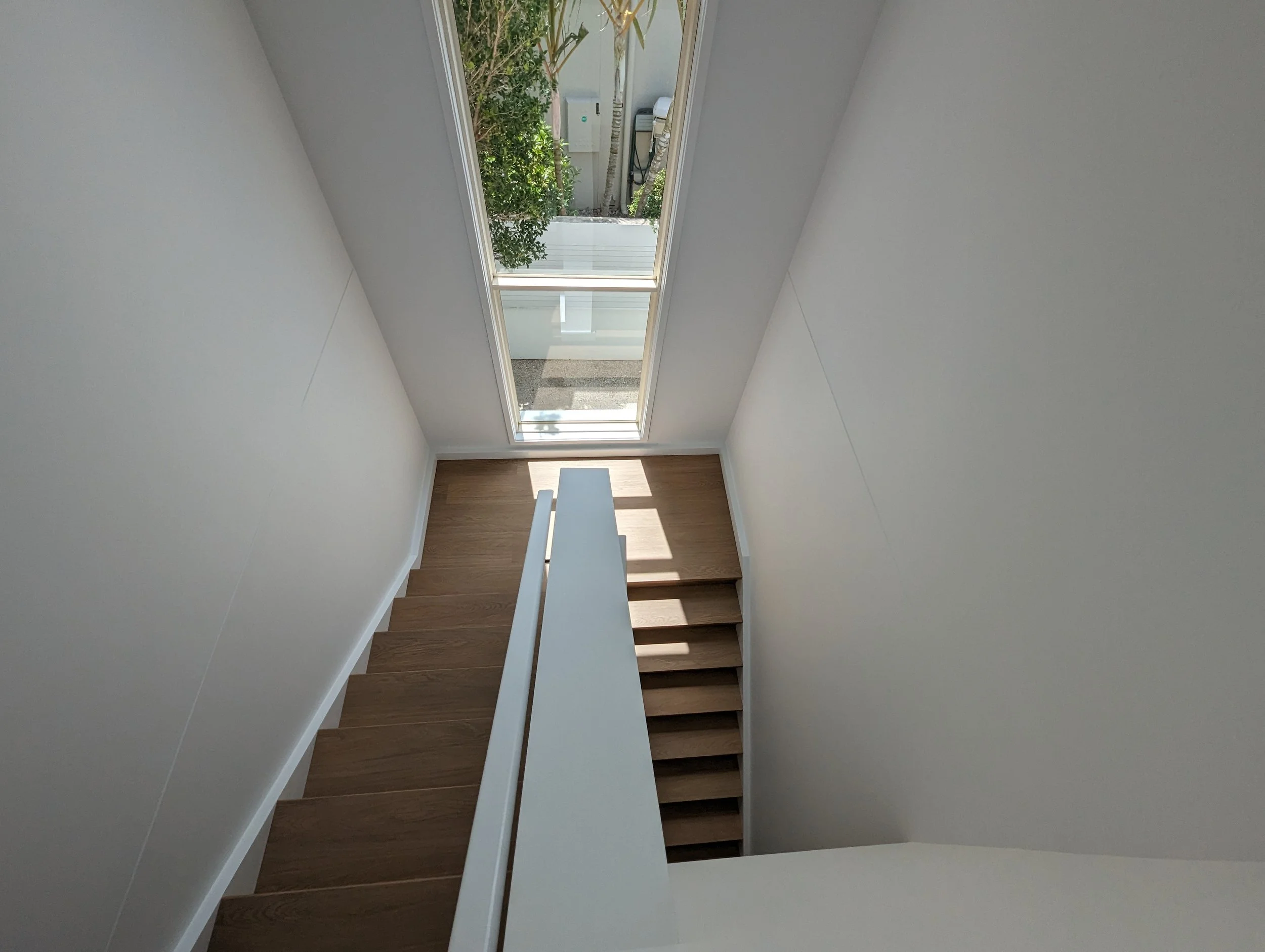 Interior view of a modern staircase with wooden steps, white walls, and a large glass door at the top letting in natural light, revealing a small outdoor area with green foliage.