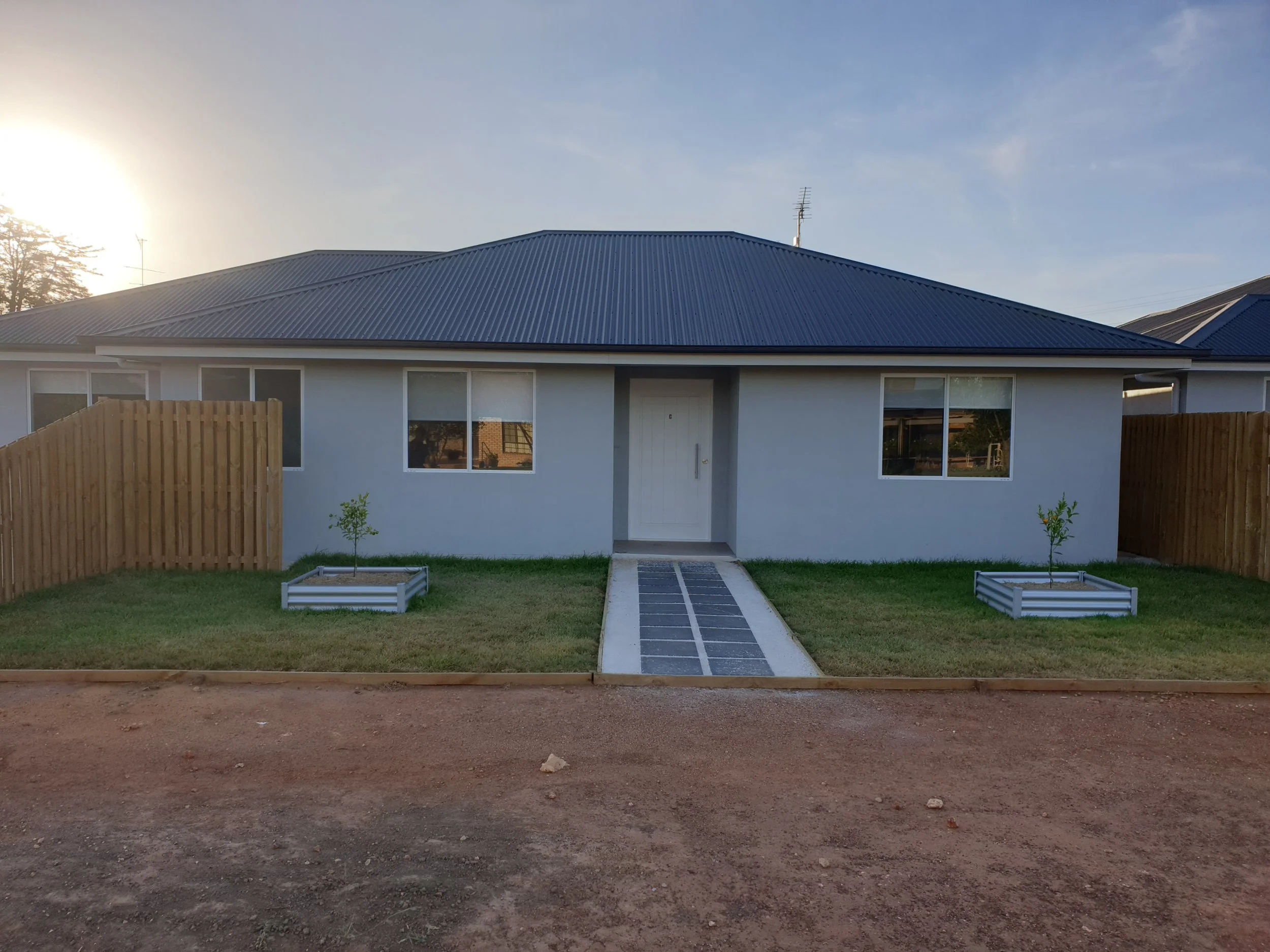 Front view of a modern, small house with a blue metal roof, two windows, a white door, and two small trees in planter boxes on a lawn, with a dirt road in the foreground and a fence on each side.