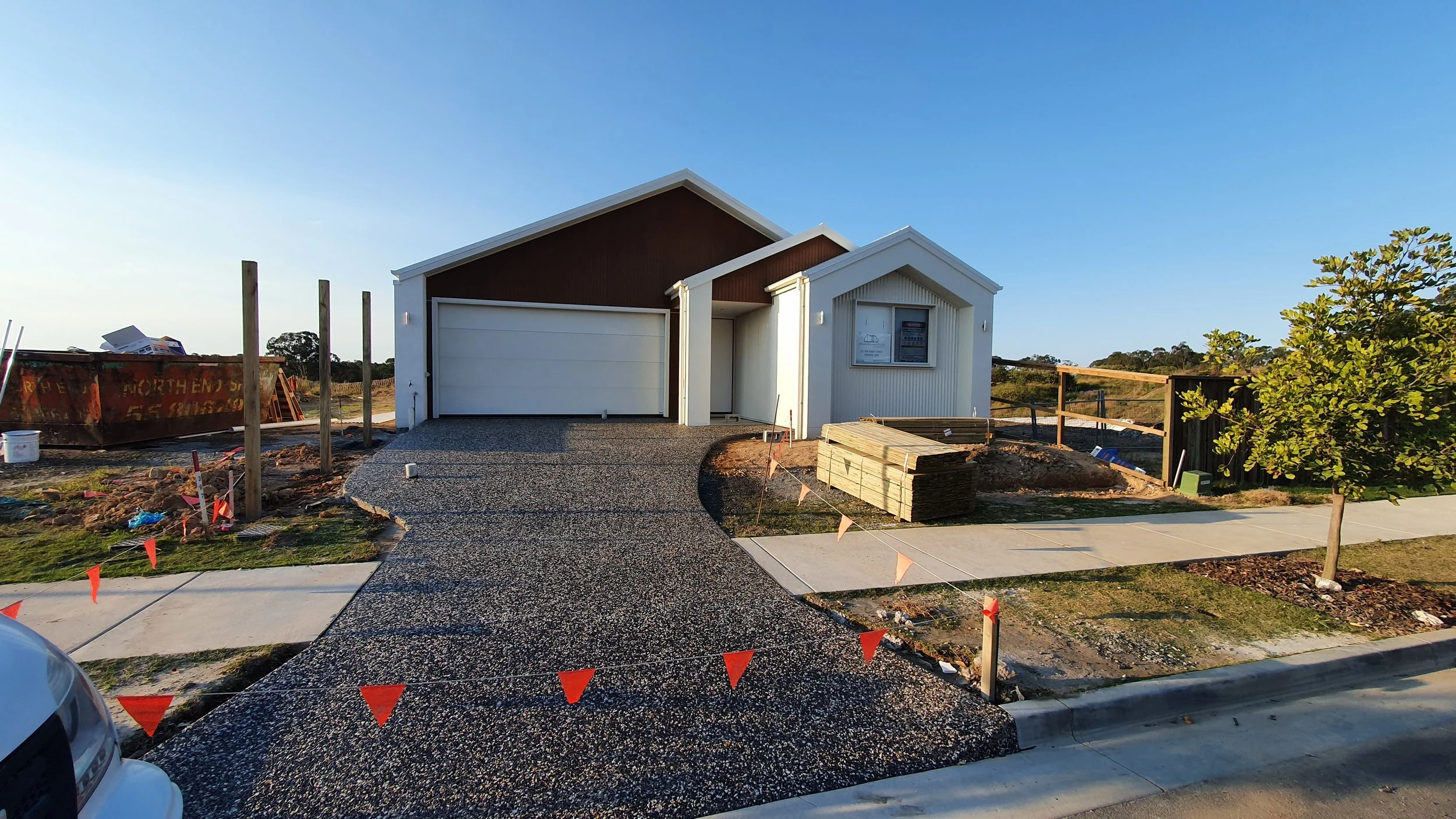 Newly constructed modern house with a curved pebble driveway, surrounded by a construction site with building materials and safety flags, under a clear blue sky.