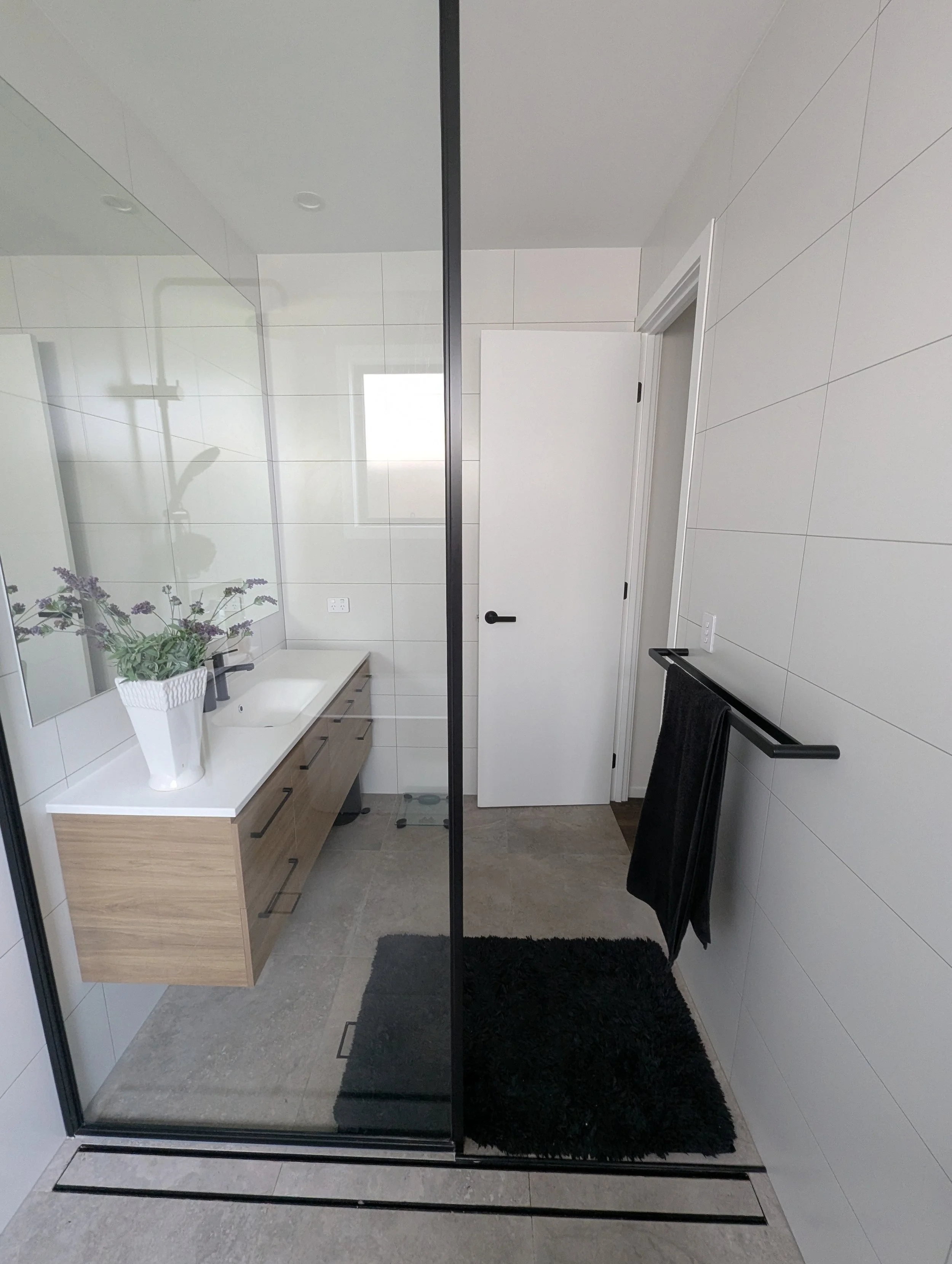 Modern bathroom with white walls, a wooden vanity with a white countertop, a potted plant, a black towel rack with a black towel, a black rug, and a glass shower door.