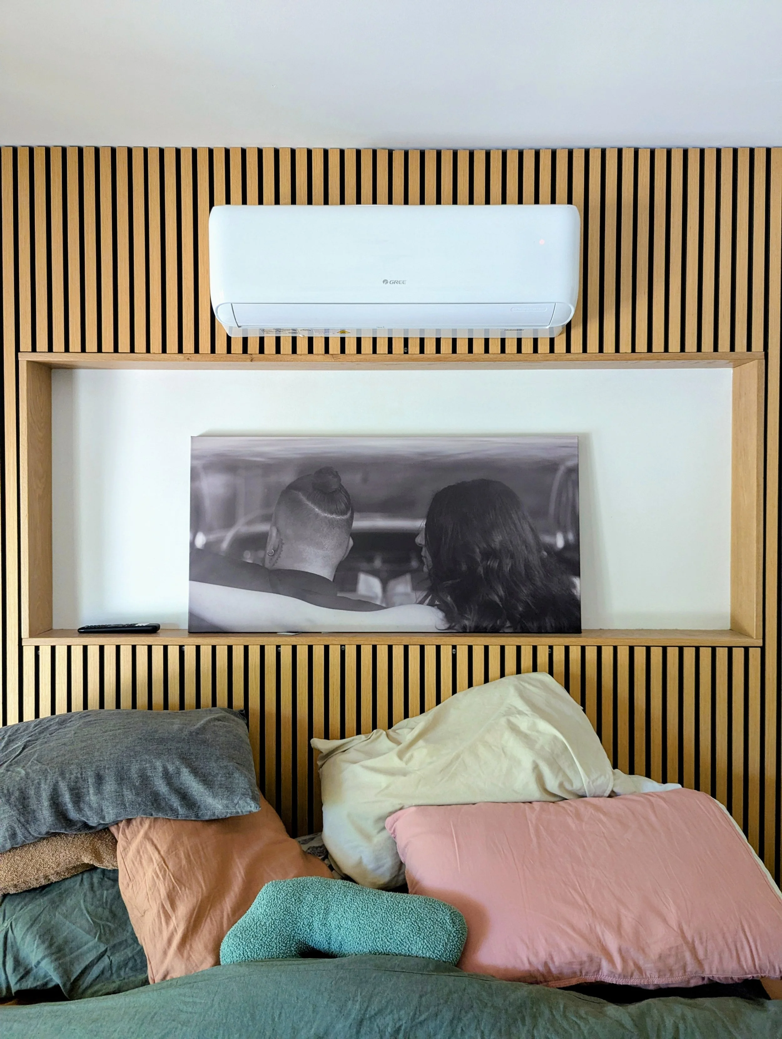 A bedroom with a bed covered in multiple pillows of different colors, a framed black-and-white photo of two people on the wall, a remote control on the frame, and an air conditioning unit above the photo.