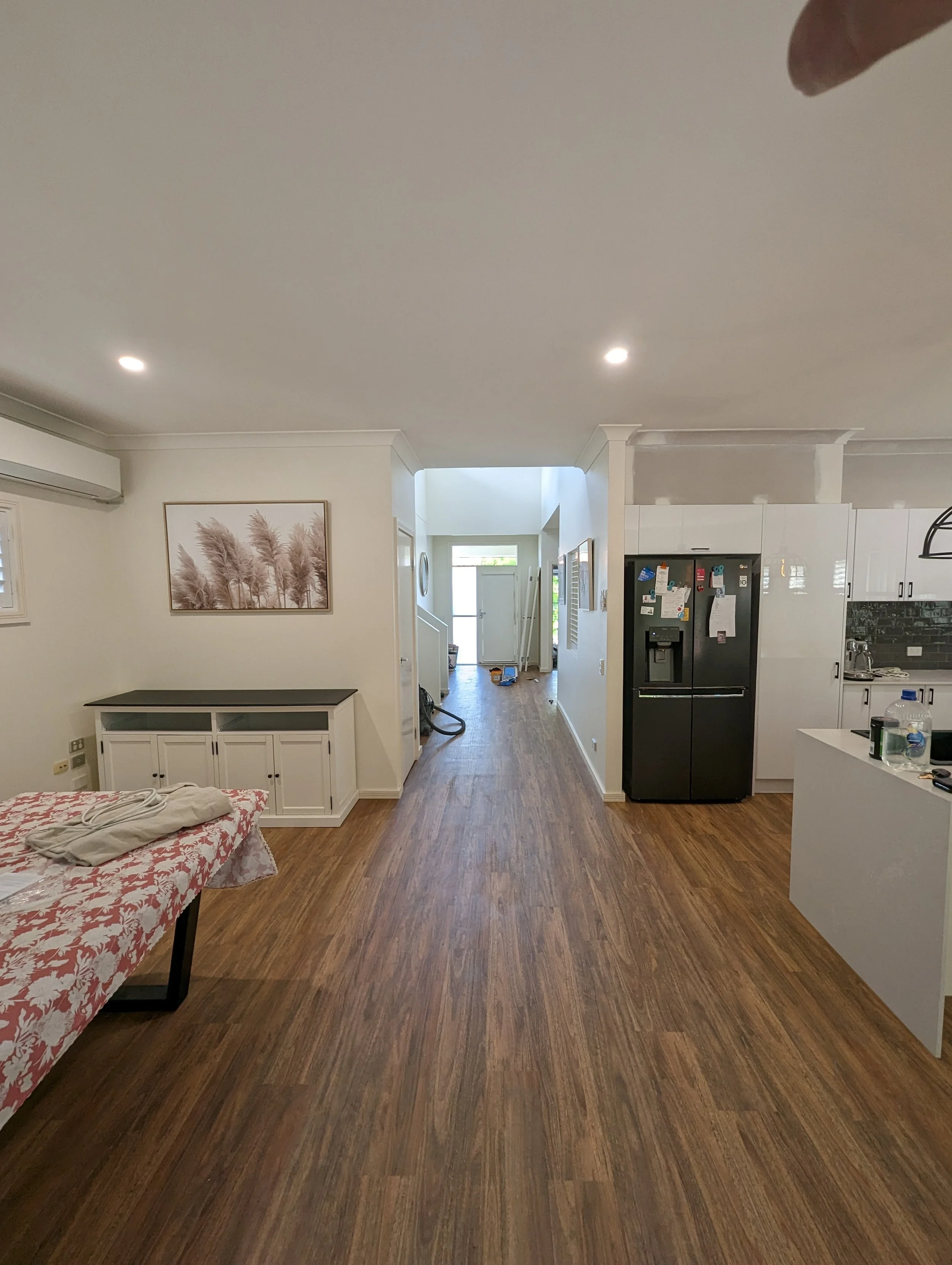 Interior view of a living space with wooden flooring, a black refrigerator, white cabinetry, a kitchen area with black tile backsplash, and a hallway leading to an entry door, with a hanging light fixture and wall art.