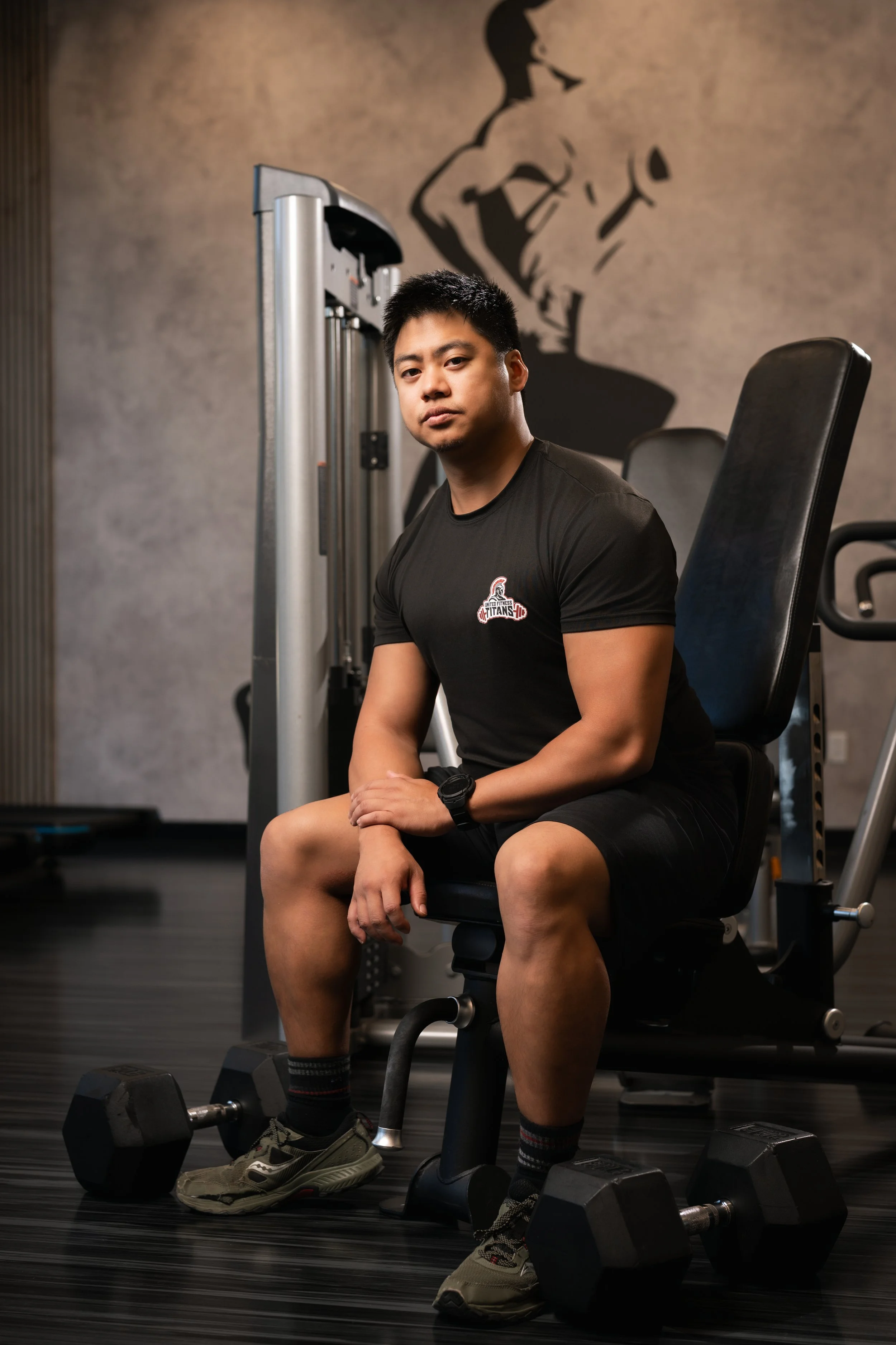 A young man sitting on a gym chair in a fitness center, surrounded by dumbbells, with a large fitness-themed mural on the wall behind him.